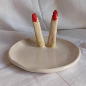 May include: White ceramic ring dish with two sculpted fingers with red nail polish.