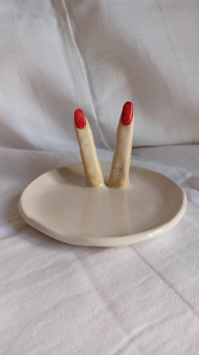 May include: White ceramic ring dish with two sculpted fingers with red nail polish.