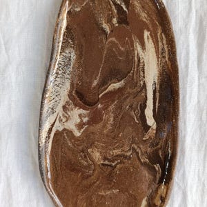 Può includere: Un vassoio in ceramica fatto a mano, di forma organica, con una glassa marmorizzata marrone e crema. Il vassoio ha una finitura lucida ed è posto su uno sfondo di lino bianco. Questo pezzo decorativo è adatto per servire o esporre.