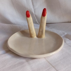 May include: A white ceramic ring dish with two fingers sticking up. The fingers are painted a light skin tone and have red nail polish.