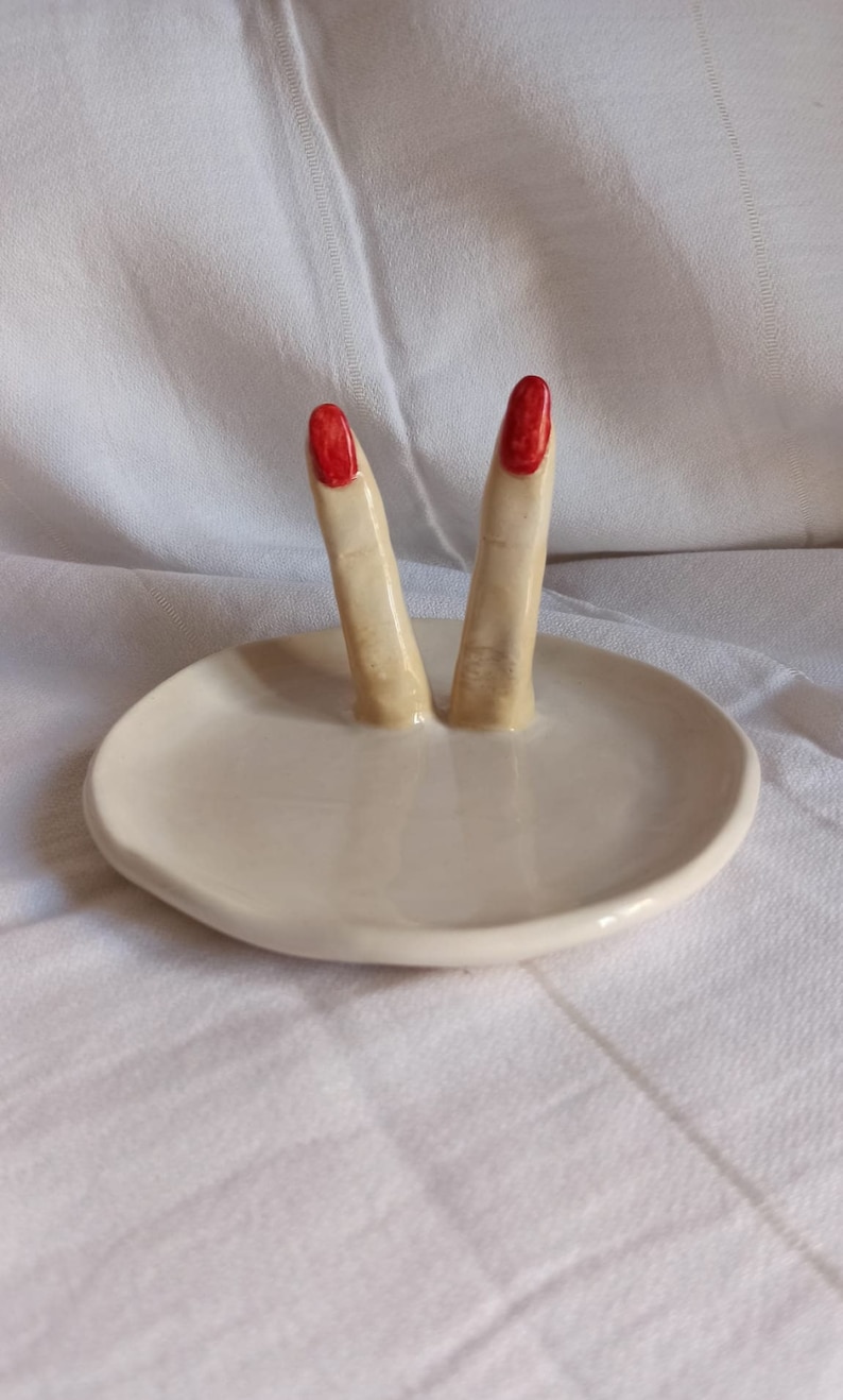 May include: A white ceramic ring dish with two fingers sticking up. The fingers are painted a light skin tone and have red nail polish.