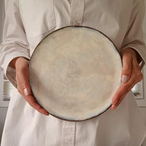 Può includere: Un piatto rotondo in ceramica bianco sporco con bordo marrone scuro. Il piatto ha una superficie marmorizzata e strutturata. Il piatto è tenuto da una persona che indossa una camicia bianca. È un articolo per la cucina.