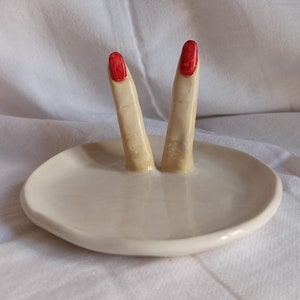 May include: White ceramic ring dish with two fingers sticking up, painted with red nail polish.