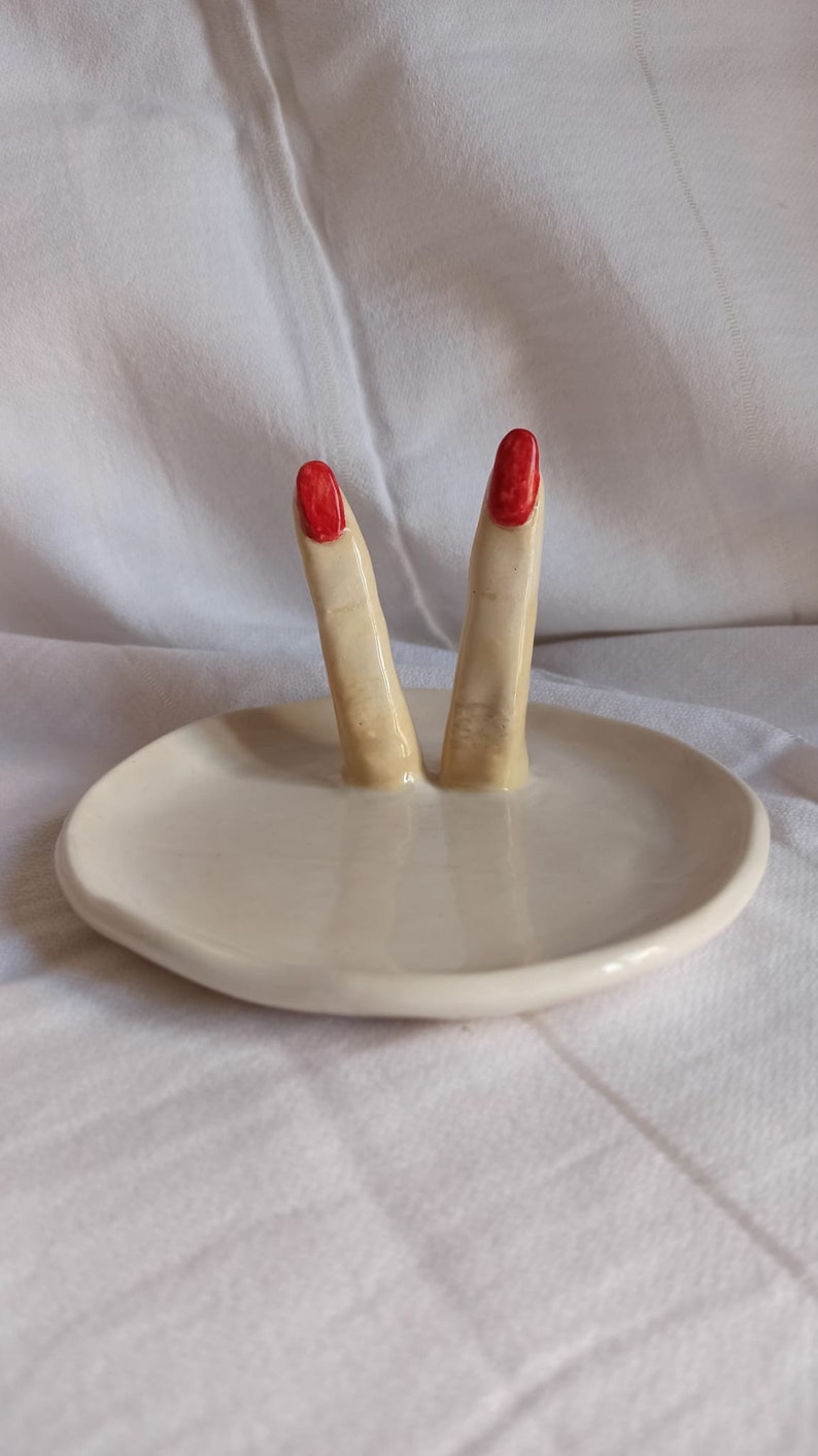 May include: White ceramic ring dish with two fingers sticking up, painted with red nail polish.