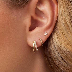 May include: Gold hoop earrings with a single row of small clear cubic zirconia stones. The earrings are shown in a person's ear with a smaller gold stud earring with three clear cubic zirconia stones.