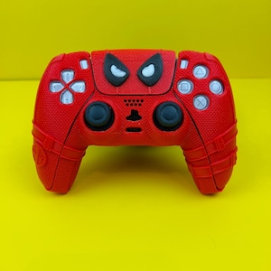 May include: A red PlayStation 5 controller with a Spider-Man design. The controller has a black and white Spider-Man face on the front.