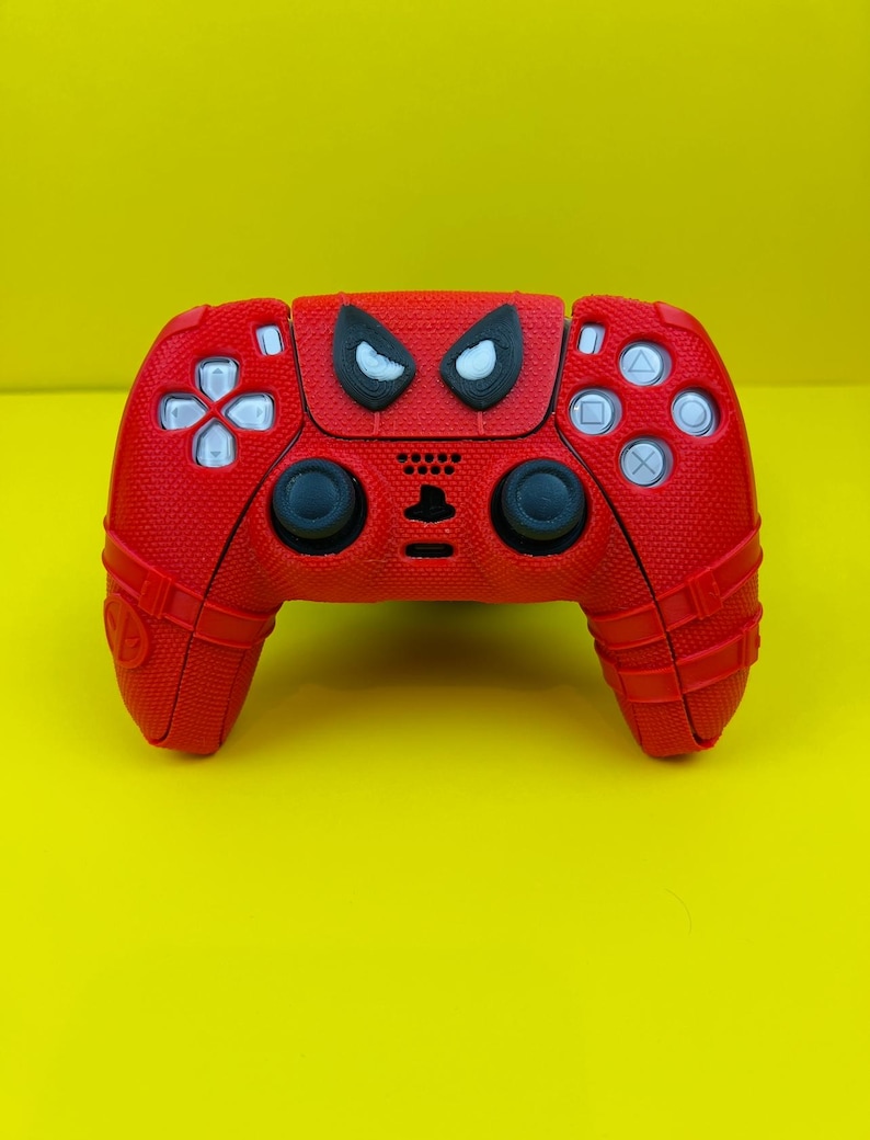 May include: A red PlayStation 5 controller with a Spider-Man design. The controller has a black and white Spider-Man face on the front.