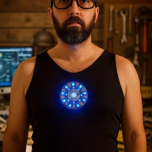 May include: A black vest top featuring a glowing blue arc reactor design on the chest. The arc reactor is circular with a detailed pattern and emits a bright blue light. The background is a wooden wall.