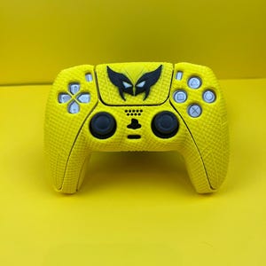 May include: A yellow PlayStation 5 controller with a black Wolverine logo on the front. The controller has a textured surface.
