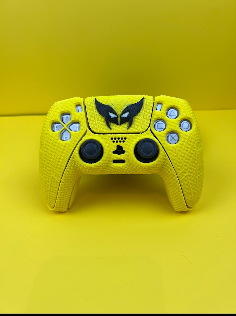 May include: A yellow PlayStation 5 controller with a black Wolverine logo on the front. The controller has a textured surface.
