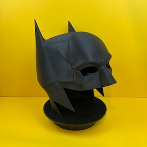 Puede incluir: Una máscara de Batman de plástico negro con una textura en relieve en la superficie. La máscara está sobre un soporte circular negro.