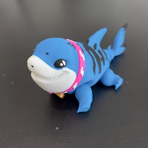 Puede incluir: Una figurita de tiburón azul y negro con un collar rosa y un dije dorado. El tiburón tiene una boca blanca y ojos negros.