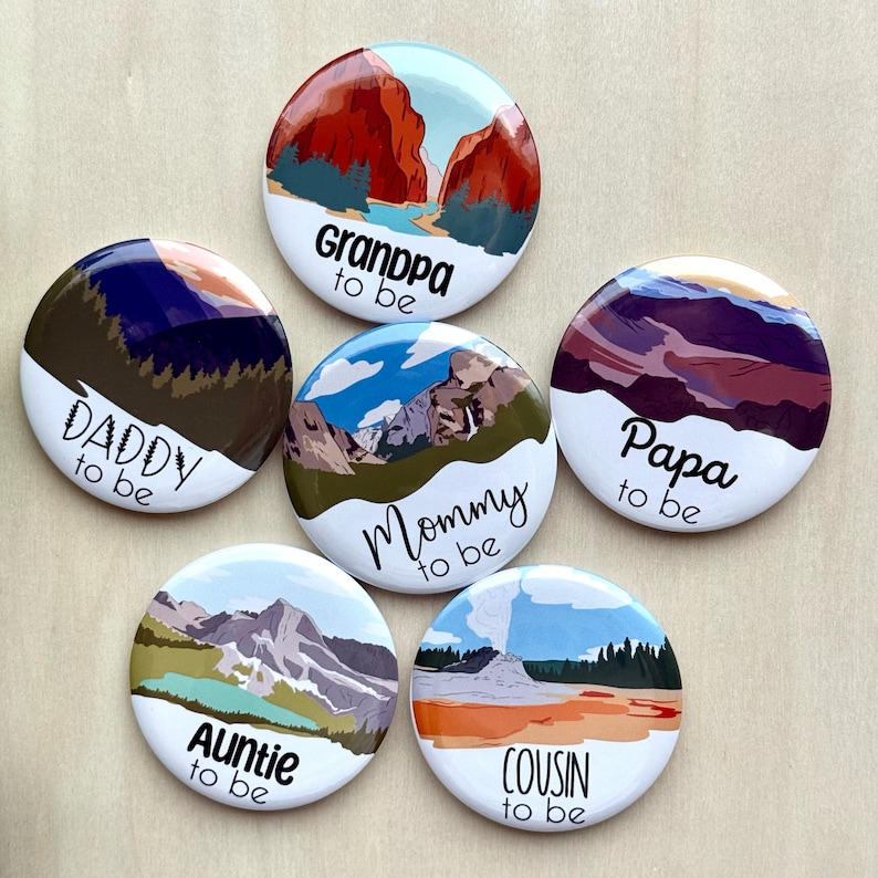 May include: A collection of six round buttons with landscape illustrations and the phrases "Daddy to be", "Grandpa to be", "Mommy to be", "Papa to be", "Auntie to be", and "Cousin to be" in black script. Each button features a different scenic design.