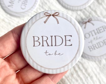 May include: A round, white button with the word "BRIDE" in brown script, and "to be" in a smaller font. The button has a striped border and a brown bow graphic. Other buttons with wedding-related text are visible.