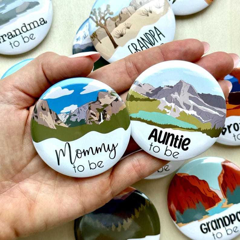 May include: A collection of white, round buttons with landscape illustrations and text. The buttons feature mountain scenes and phrases like "Mommy to be" and "Auntie to be" in black script. The buttons are held in a hand.