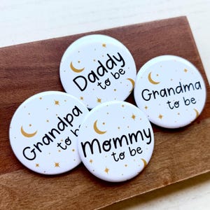 May include: Four white round buttons with gold crescent moons and stars. The buttons read "Daddy to be", "Grandma to be", "Mommy to be", and "Grandpa to be" in black text. The buttons are on a brown wooden surface.