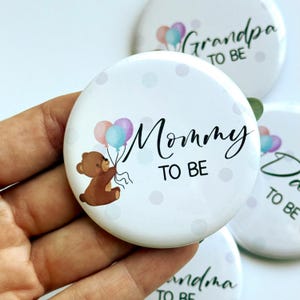 以下が含まれることがあります： 黒い筆記体で「Mommy TO BE」と書かれた白いボタン。茶色のテディベアが風船を持っています。ボタンは水玉模様で、セットの一部です。