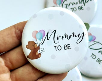 May include: White button with the text "Mommy TO BE" in black script, featuring a brown teddy bear holding balloons. The button has a polka dot pattern and is part of a set.