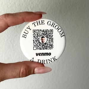 Könnte beinhalten: Runder weißer Knopf mit schwarzem Text, der "Buy the Groom a Drink" lautet. Ein QR-Code mit dem Gesicht eines Mannes in der Mitte ist vom Text umgeben. Der Text "venmo" befindet sich unter dem QR-Code.