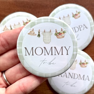 May include: Round button with a green and white gingham border. The center features a clothesline with a baby onesie, hat, and basket of fruit. The text "MOMMY to be" is displayed in a script font.