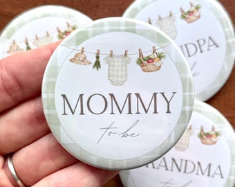 May include: Round button with a green and white gingham border. The center features a clothesline with a baby onesie, hat, and basket of fruit. The text "MOMMY to be" is displayed in a script font.