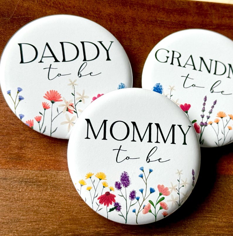 May include: Three white button pins with floral designs and black text. The text on the pins reads "Daddy to be", "Mommy to be", and "Grandma to be".