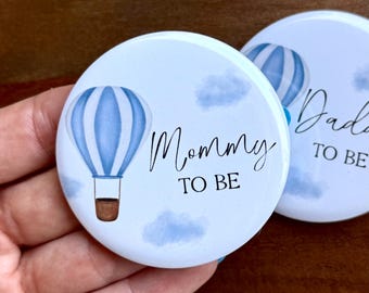 May include: Two round buttons with a light blue and white hot air balloon design. One button says "Mommy TO BE" and the other says "Daddy TO BE". The buttons are white with blue clouds in the background.