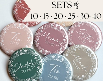 May include: A collection of round pin-back buttons in various muted colors including dusty rose, sage green, blue, and beige. Each button features a white lace-style border and white text such as Mommy to be, Daddy to be, Tia to be, Tio to be, Sister to be, and Abuela to be.