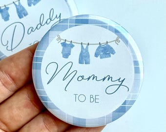 May include: Round button with a blue and white plaid border. The center has the words "Mommy TO BE" in cursive, with a clothesline above it holding three blue baby clothes. Another button with the word "Daddy" is visible.