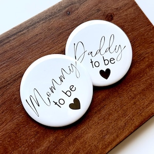 Pode incluir: Dois botões redondos brancos com texto preto que diz "Mommy to be" e "Daddy to be". Cada botão tem um coração preto. Os botões estão sobre uma superfície de madeira castanha.