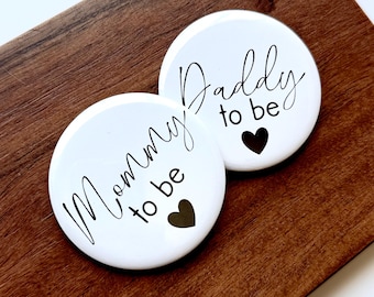 May include: Two white round buttons with black text that reads "Mommy to be" and "Daddy to be". Each button has a black heart. The buttons are on a brown wooden surface.
