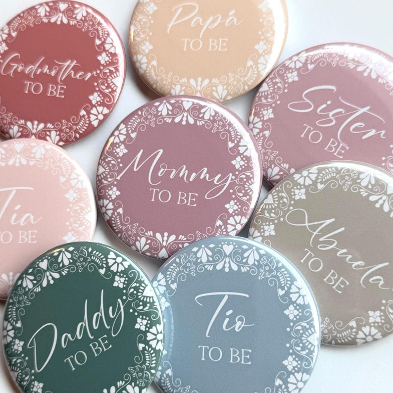 May include: A set of nine round buttons with floral designs and white text. The text on the buttons reads "Godmother to Be", "Tia to Be", "Pap&aacute; to Be", "Sister to Be", "Mommy to Be", "Abuela to Be", "Daddy to Be", "Tio to Be".