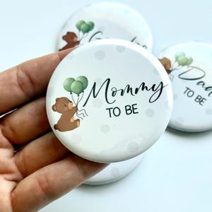 以下が含まれることがあります： 「Mommy TO BE」の黒い筆記体文字が入った白い丸いボタン。茶色のテディベアが緑色の風船を持っています。ボタンには水玉模様があり、手で持たれています。