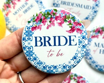 May include: White and blue floral design button with the text "BRIDE to be" in navy blue and red. The button has a floral border with pink flowers and green leaves. Other buttons with text like "Bridesmaid" are visible.