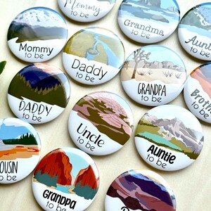 May include: A collection of round, white buttons with landscape illustrations and text. The buttons feature phrases like "Mommy to be," "Daddy to be," "Grandpa to be," and "Auntie to be." The illustrations depict mountains, rivers, and desert scenes.