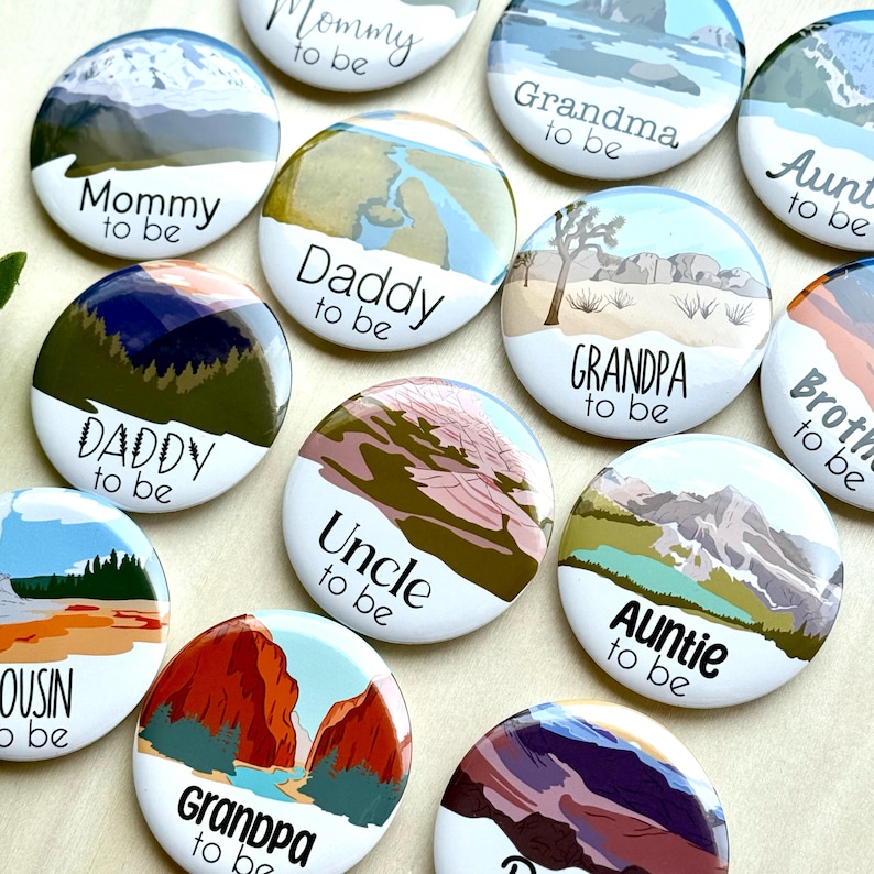 May include: A collection of round, white buttons with landscape illustrations and text. The buttons feature phrases like "Mommy to be," "Daddy to be," "Grandpa to be," and "Auntie to be." The illustrations depict mountains, rivers, and desert scenes.