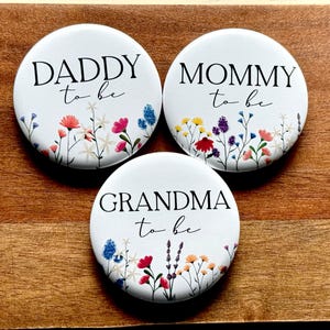 May include: Three white round buttons with floral designs and black text. The buttons read "Daddy to be", "Mommy to be", and "Grandma to be".