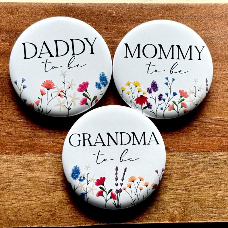 May include: Three white round buttons with floral designs and black text. The buttons read "Daddy to be", "Mommy to be", and "Grandma to be".