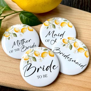Puede incluir: Cuatro insignias redondas blancas para la fiesta de bodas con diseños de limones y flores. El texto incluye "Mother of the Bride", "Maid of Honor", "Bridesmaid" y "Bride to Be". Un limón y vegetación en el fondo.