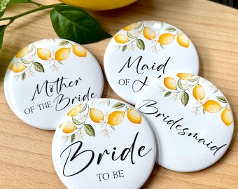 May include: Four white, round bridal party badges with lemon and floral designs. Text includes "Mother of the Bride", "Maid of Honor", "Bridesmaid", and "Bride to Be". A lemon and greenery are in the background.
