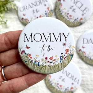 Puede incluir: Botón redondo blanco con el texto "MOMMY to be" en escritura negra. El botón está decorado con un diseño floral con flores rojas, azules y amarillas, mariposas y libélulas. Se ven otros botones con diseños similares.