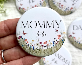 May include: White round button with the text "MOMMY to be" in black script. The button is decorated with a floral design featuring red, blue, and yellow flowers, butterflies, and dragonflies. Other buttons with similar designs are visible.