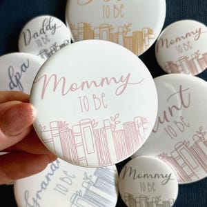 Può includere: Un set di nove spille a bottone bianche con testo rosa e dorato e illustrazioni di libri. Il testo sulle spille dice "Mommy to be", "Daddy to be", "Aunt to be", "Grandma to be" e "Grandpa".