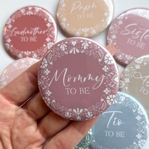 May include: A set of pink, peach, and blue round buttons with white floral designs and white text. The text on the buttons reads "Godmother to Be", "Papa to Be", "Sister to Be", "Mommy to Be", "Tio to Be", and "Auntie to Be".
