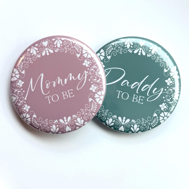 May include: Two round buttons with white floral designs and the text "Mommy to Be" on a pink button and "Daddy to Be" on a teal button.
