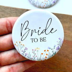 May include: White round button with the text "Bride to Be" in black script. The button is decorated with a border of colorful wildflowers.
