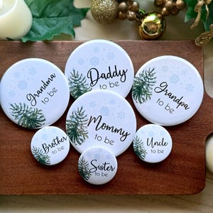 May include: Set of 8 round white buttons with a wintery design. Each button features a pine tree and snowflakes with text that reads "Grandma to be", "Brother to be", "Daddy to be", "Mommy to be", "Sister to be", "Grandpa to be", "Uncle to be".