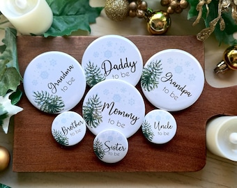 May include: Set of 8 round white buttons with a wintery design. Each button features a pine tree and snowflakes with text that reads "Grandma to be", "Brother to be", "Daddy to be", "Mommy to be", "Sister to be", "Grandpa to be", "Uncle to be".