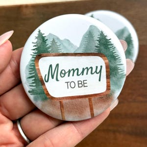May include: Two round white buttons with a watercolor design. The buttons feature a mountain and forest scene with the text "Mommy TO BE" in green script on a wooden sign. The buttons are held in a hand.