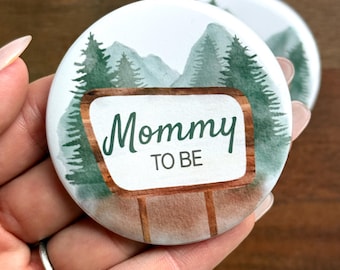 May include: Two round white buttons with a watercolor design. The buttons feature a mountain and forest scene with the text "Mommy TO BE" in green script on a wooden sign. The buttons are held in a hand.